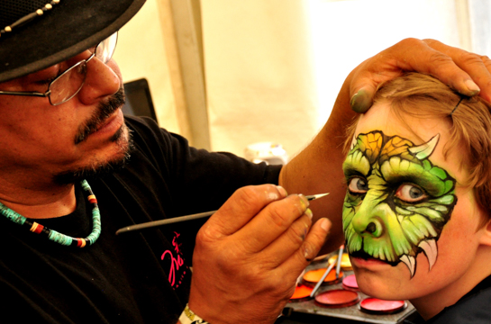 Mark Reid painting a lizzard mask on a young person who is looking at the camera